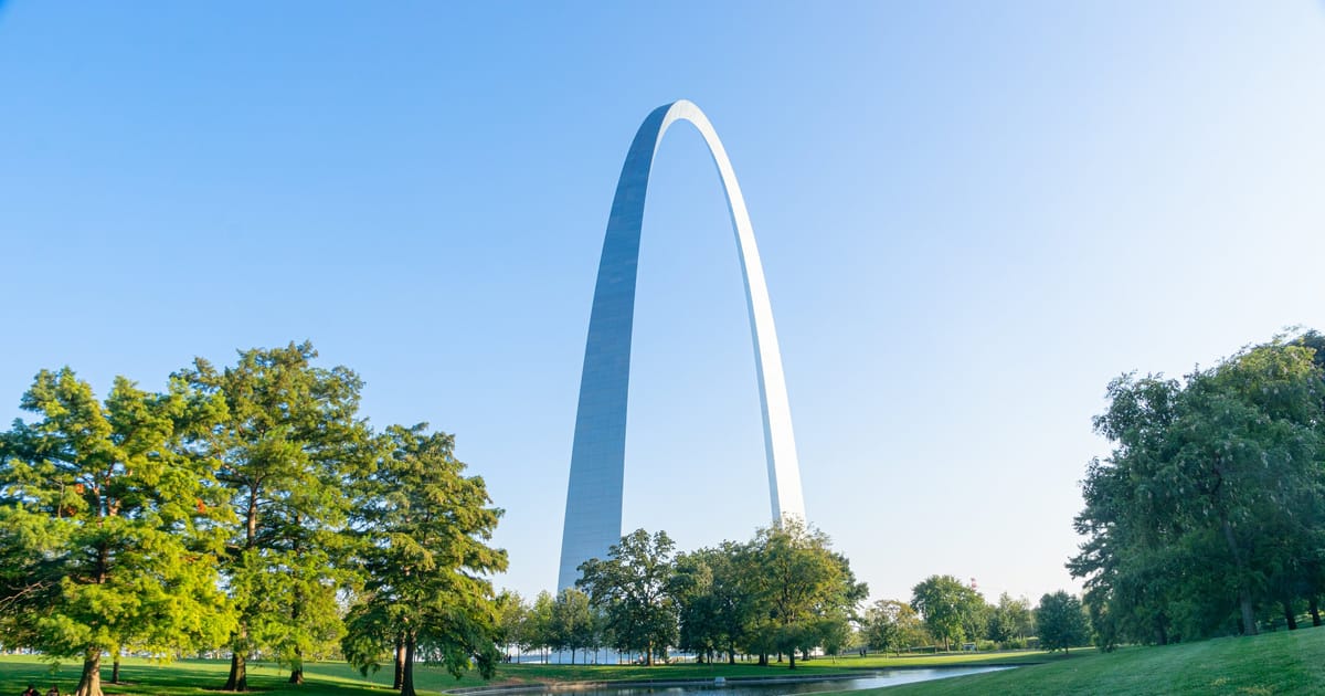 The iconic Gateway Arch in St. Louis rises dramatically against a clear blue sky, reflected in a calm reflecting pool surrounded by lush green parkland. Warm morning light illuminates the stainless steel structure and verdant trees, creating a…