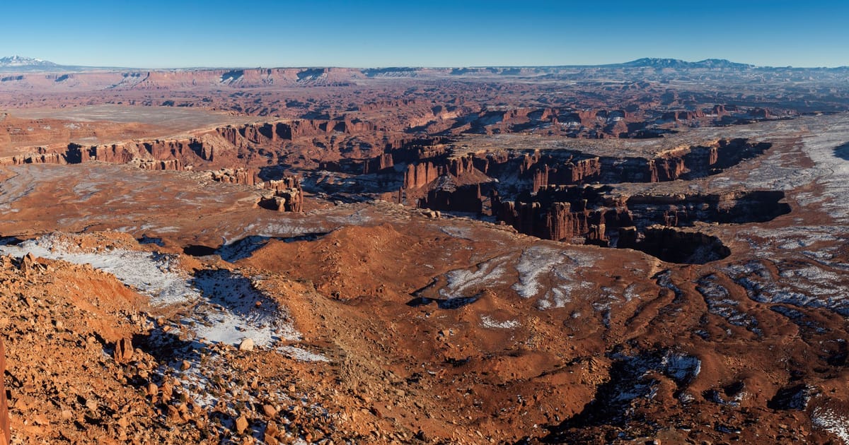 The Green River overlook in Canyonlands National Park, showing the river as a pale ribbon carving through deep red canyon walls and layered sandstone terraces, as seen from the Island in the Sky mesa under afternoon light.