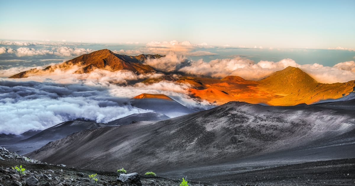 A dramatic aerial view of Haleakalā volcanic crater on Maui, Hawaii, with cinder cones bathed in warm golden-orange sunrise light surrounded by a sea of clouds. The stark dark volcanic terrain in the foreground contrasts with vibrant illuminated…