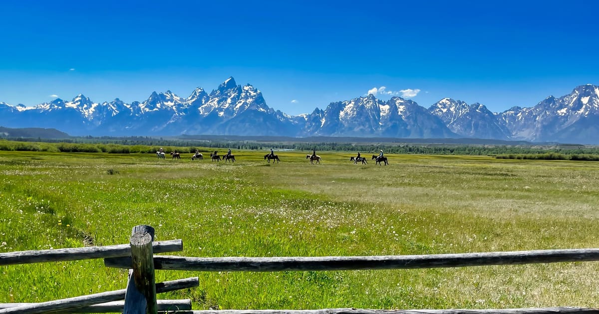 A sweeping summer meadow in Grand Teton National Park features a group of horseback riders crossing a vast green valley, with the iconic jagged, snow-capped Teton Range dramatically filling the horizon under a brilliant blue sky. A weathered…
