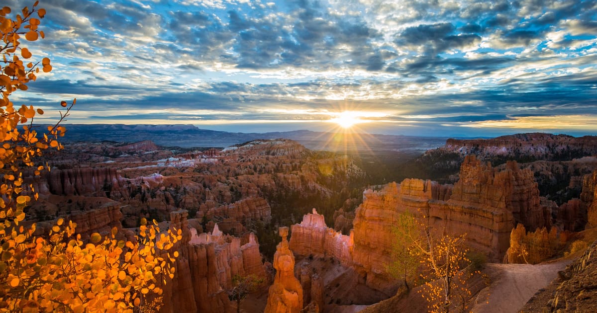 Visitors at Inspiration Point overlook in Bryce Canyon National Park, gazing across the vast Natural Bridge amphitheater filled with thousands of hoodoo spires at sunset. The formations glow in deep orange and red as long shadows stretch across the canyon floor.