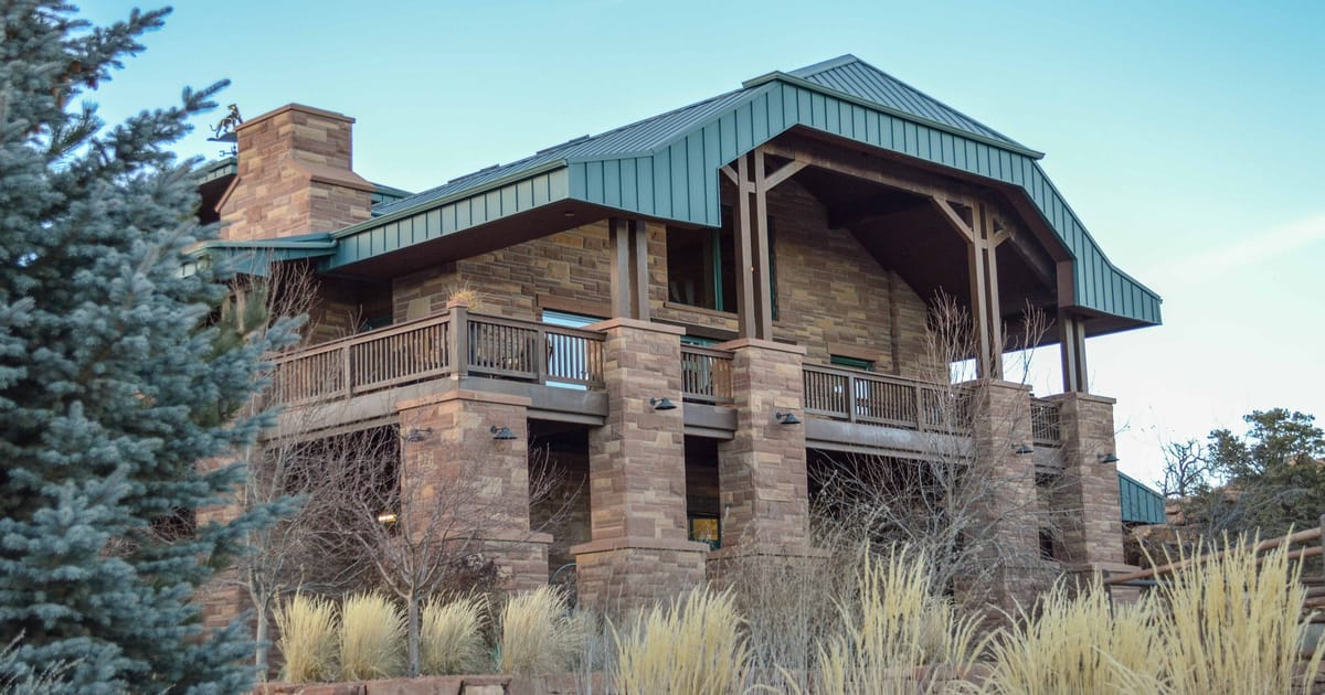 A multi-story rustic lodge building constructed of layered sandstone and warm-toned stone masonry with a green standing-seam metal roof, surrounded by ornamental grasses and sparse desert vegetation under a clear blue sky. The mood is quiet and subdu