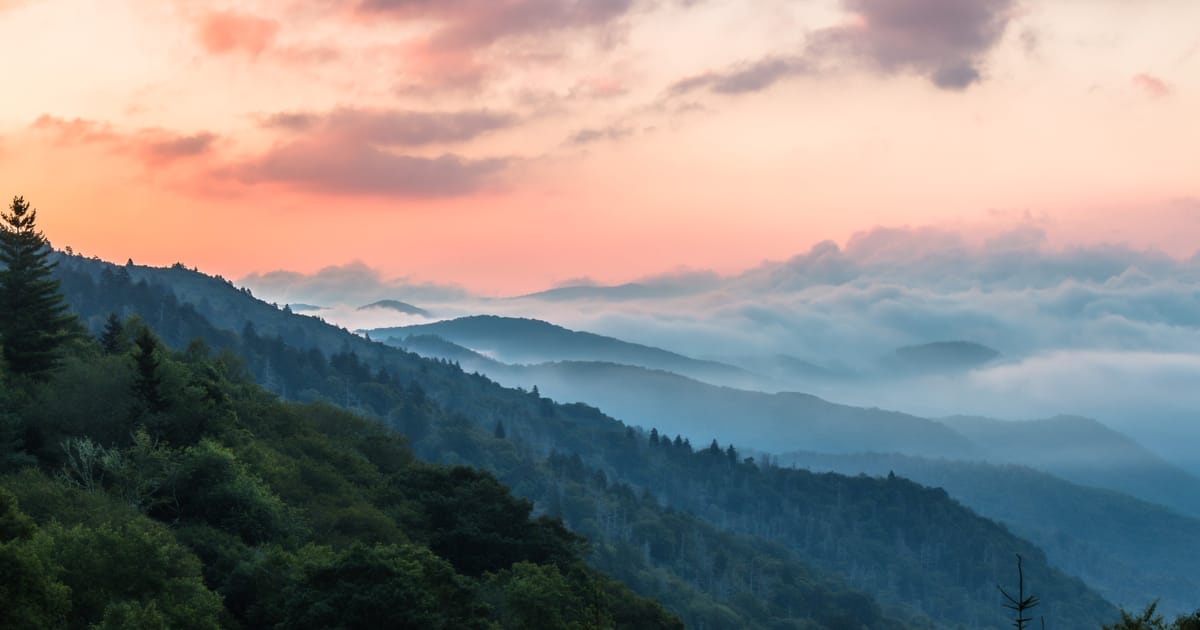 A sweeping panoramic view of layered, mist-filled mountain valleys covered in dense deciduous and evergreen forest, captured at sunrise or sunset with a dramatic pink and orange sky punctuated by soft clouds and flowing fog banks between the…