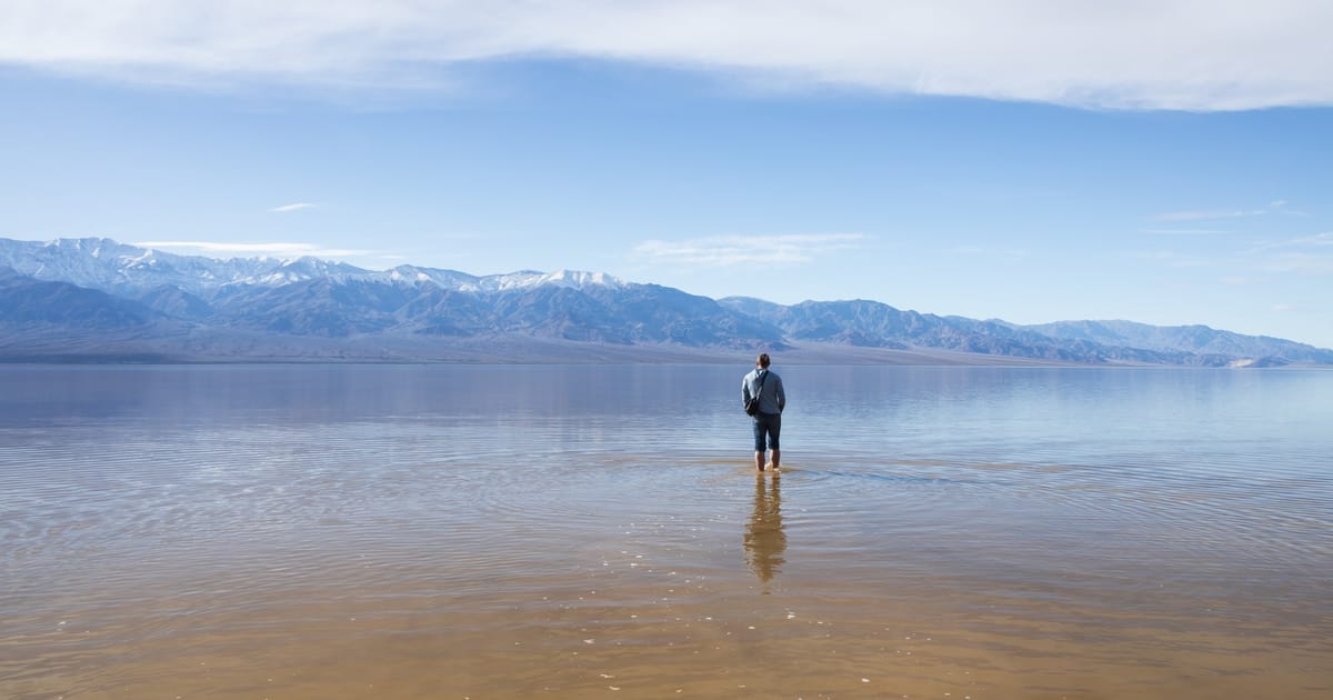 A lone figure wades through the shallow, mirror-like waters of a flooded desert basin — likely Badwater Basin in Death Valley National Park — with snow-dusted mountain ranges stretching across the horizon under a expansive blue sky with wispy…