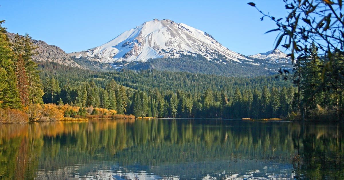 A snow-capped volcanic peak — likely Lassen Peak in Lassen Volcanic National Park, California — rises above a dense evergreen forest, with its reflection mirrored perfectly in the calm, glassy surface of Manzanita Lake. The autumn foliage adds warm…