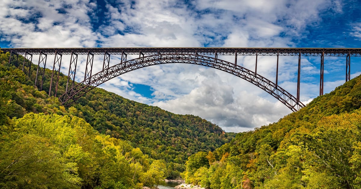 The iconic New River Gorge Bridge, a massive steel arch bridge spanning the densely forested gorge above the New River in West Virginia, photographed from below with lush green canopy and partly cloudy blue skies creating a dramatic, majestic mood.
