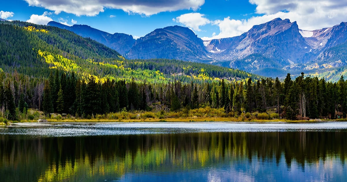 A serene alpine lake in Rocky Mountain National Park reflects the surrounding evergreen forest and rugged mountain peaks under a partly cloudy blue sky. The scene captures early autumn foliage with golden aspen trees dotting the hillsides, creating…