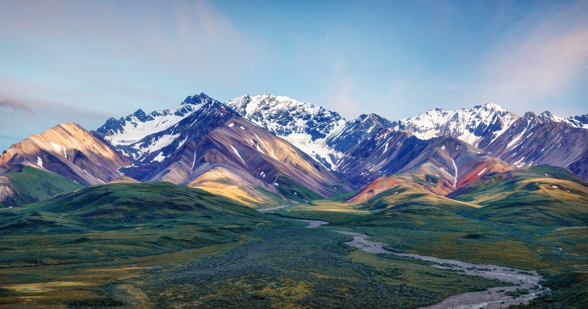 A sweeping panoramic view of the Alaska Range in Denali National Park, featuring snow-capped peaks with colorful mineral-streaked slopes in warm amber, rust, and olive tones, above a braided glacial river winding through lush green tundra valleys.…