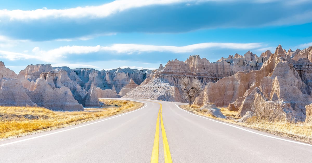 A two-lane highway with double yellow center lines winds through the dramatic eroded buttes and spires of Badlands National Park, South Dakota, under a partly cloudy blue sky. The scene has an open, inviting mood with warm golden grasses flanking…