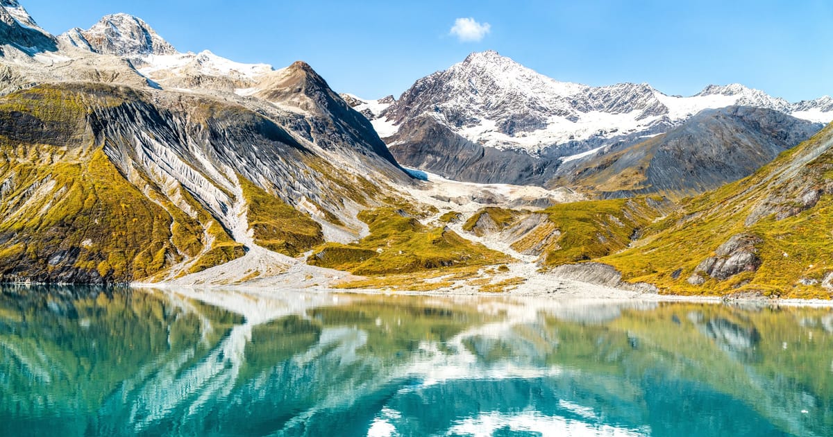 A stunning alpine lake with vivid turquoise water perfectly mirrors snow-capped mountain peaks and glaciers in calm, glassy conditions. Bright midday light illuminates golden-green tundra slopes, rocky moraines, and dramatic glaciated summits under…