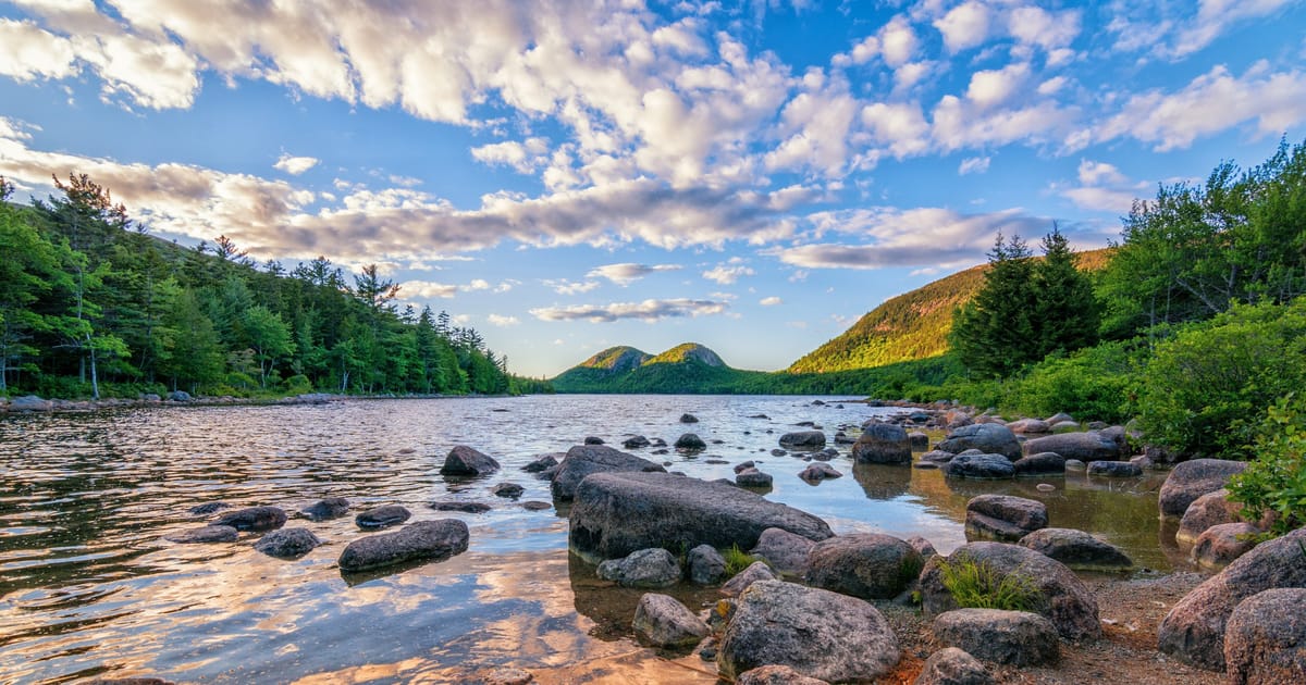 Jordan Pond in Acadia National Park, Maine, featuring smooth glacial boulders along the shoreline with the iconic Bubbles mountains rising in the background, bathed in warm golden-hour light against a dramatic blue sky filled with altocumulus clouds.