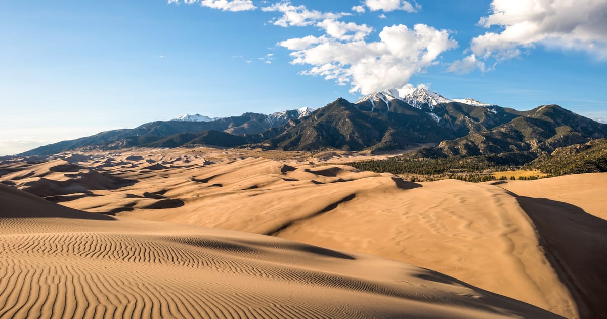Great Sand Dunes National Park in Colorado showcases towering rippled sand dunes in the foreground with snow-capped Sangre de Cristo Mountains rising dramatically in the background. Warm golden sunlight illuminates the dune ridges, creating striking…