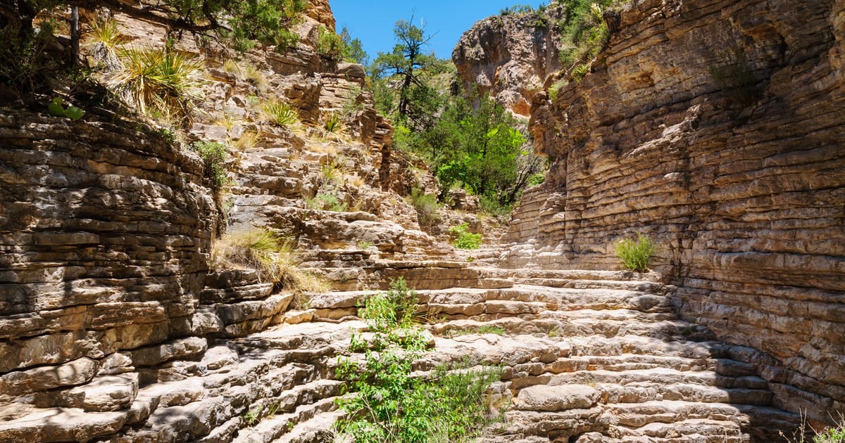 A natural stone staircase of layered limestone formations winds upward through a narrow canyon corridor, flanked by stratified rock walls with desert vegetation. Bright midday sunlight illuminates the warm tan and golden-brown rocks against a vivid…