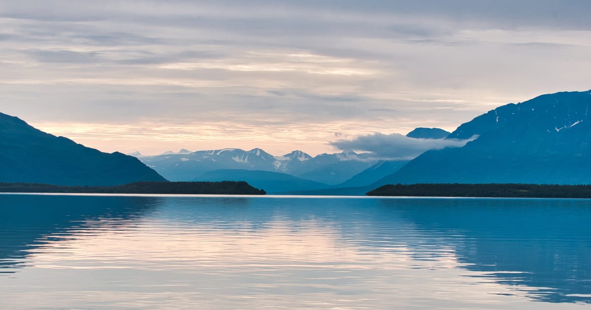 A serene glacial lake reflects soft pastel twilight hues beneath a layered overcast sky, framed by dark silhouetted mountains and snow-capped peaks receding into the misty distance. The calm, mirror-like water and cool blue tones create a tranquil,…