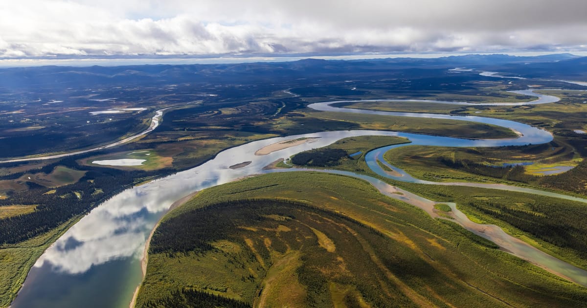 An aerial view of a vast braided and meandering river system winding through boreal forest and tundra flats, likely the Yukon or Kobuk River in Alaska, under dramatic partly cloudy skies with soft diffused light reflecting off the calm water.