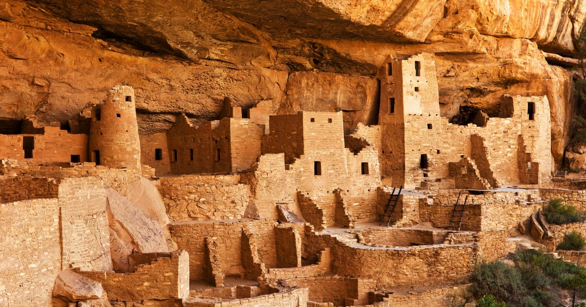 Cliff Palace at Mesa Verde National Park, an ancient Ancestral Puebloan cliff dwelling nestled beneath a massive sandstone overhang, bathed in warm golden afternoon light that accentuates the ochre and tan tones of the multi-story stone structures.