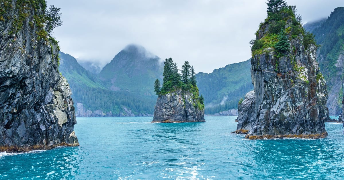 Dramatic sea stacks and rocky spires rise from vivid turquoise waters in what appears to be Kenai Fjords National Park, Alaska, with evergreen trees clinging to the summits and misty, cloud-shrouded mountains receding into the background. The…