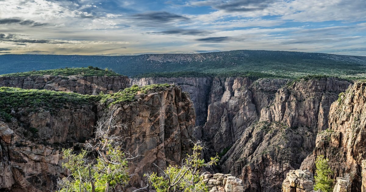 A dramatic aerial view of the Black Canyon of the Gunnison, featuring sheer dark metamorphic rock walls plunging into a deep narrow gorge, under a partially cloudy blue sky with warm afternoon light illuminating the canyon rim.