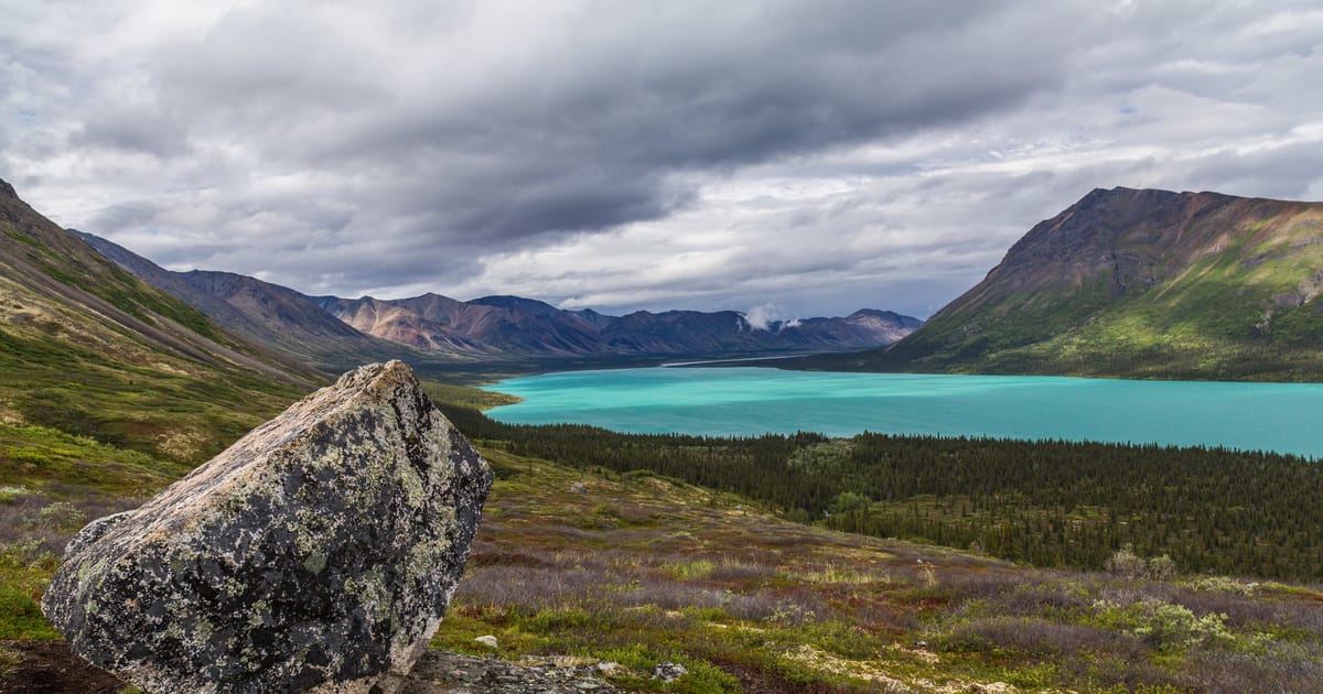 A striking glacial lake with vivid turquoise water nestled between rugged mountain ranges, framed by a large lichen-covered boulder in the foreground and a dense boreal forest below under dramatic overcast skies.