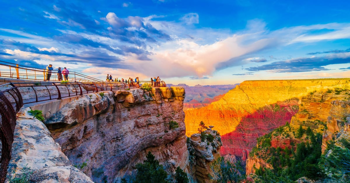 A dramatic sunset view of the Grand Canyon from Mather Point overlook on the South Rim, with warm golden and red light bathing the layered sandstone cliffs while tourists gather along the railed viewing platform.