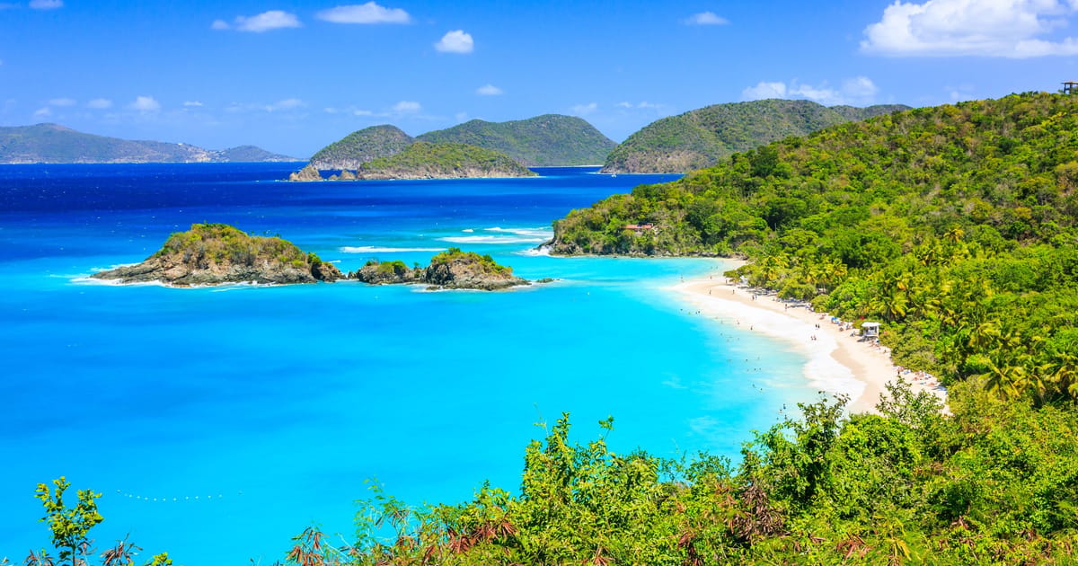 A stunning aerial view of Trunk Bay in St. John, US Virgin Islands National Park, featuring vivid turquoise and deep blue waters curving around a white sandy beach flanked by lush tropical hillsides. The scene is bathed in brilliant midday sunlight,…