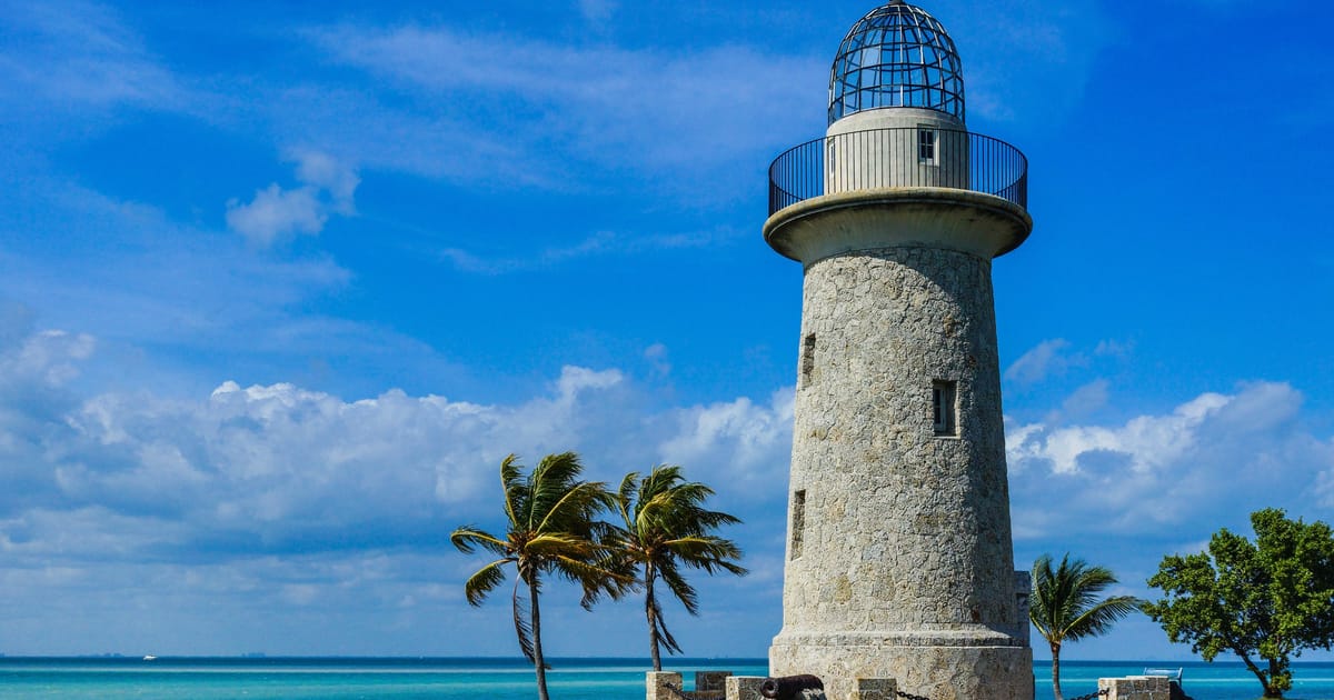 The Boca Chita Key lighthouse in Biscayne National Park stands on a small coral island surrounded by turquoise waters, with palm trees swaying in the breeze under a vibrant blue sky with scattered clouds. The historic stone tower is bathed in bright…
