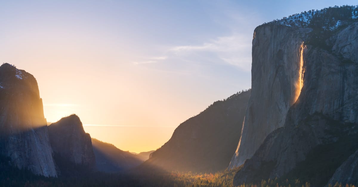 Yosemite Valley at golden hour featuring El Capitan's sheer granite face with Horsetail Fall glowing orange in the famous 'Firefall' effect, framed by forested valley floor with autumn foliage and a warm sunset illuminating misty air between canyon…