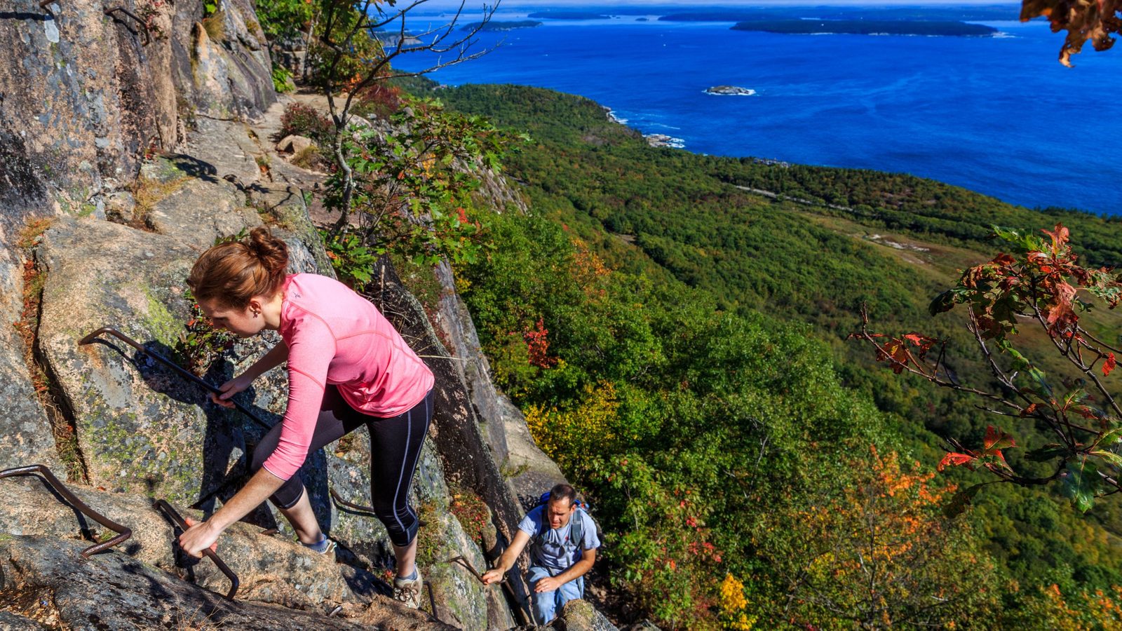 Two hikers climb a steep iron-rung ladder route bolted into exposed granite rock face in Acadia National Park, with a sweeping view of forested hillsides, co…