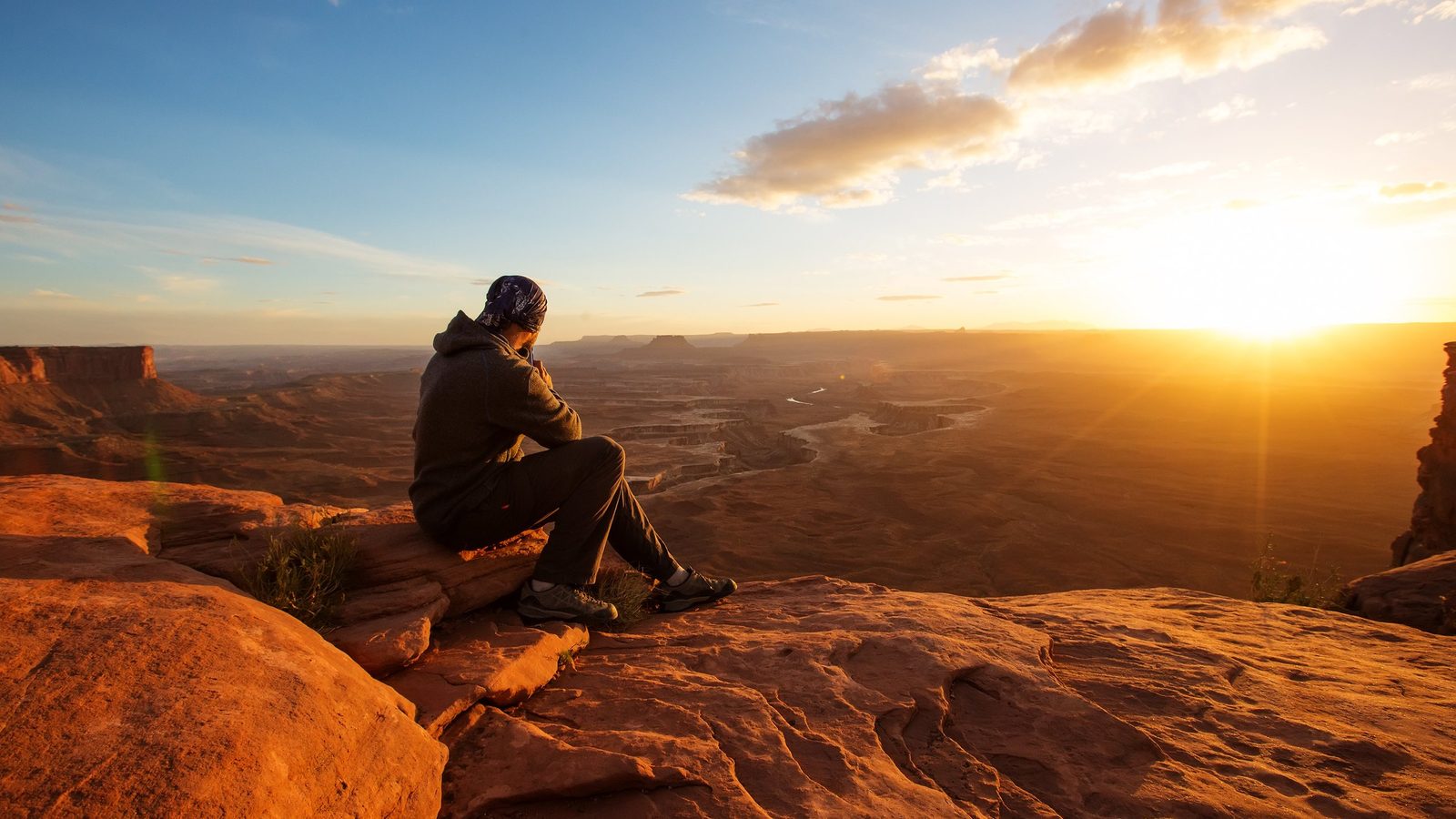 A lone hiker sits on the edge of a red sandstone cliff at Canyonlands National Park, gazing over a vast canyon landscape with a winding river below, bathed i…