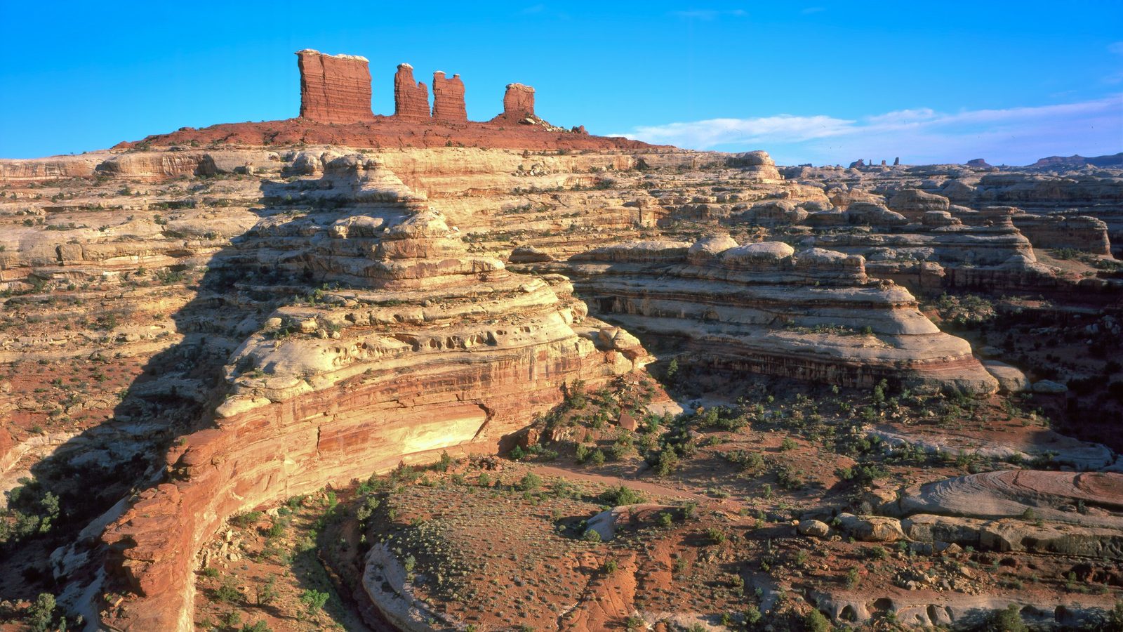 Aerial view of layered red and tan sandstone terraces in a desert canyon landscape, with three tall red rock spires on the horizon under a clear blue sky.