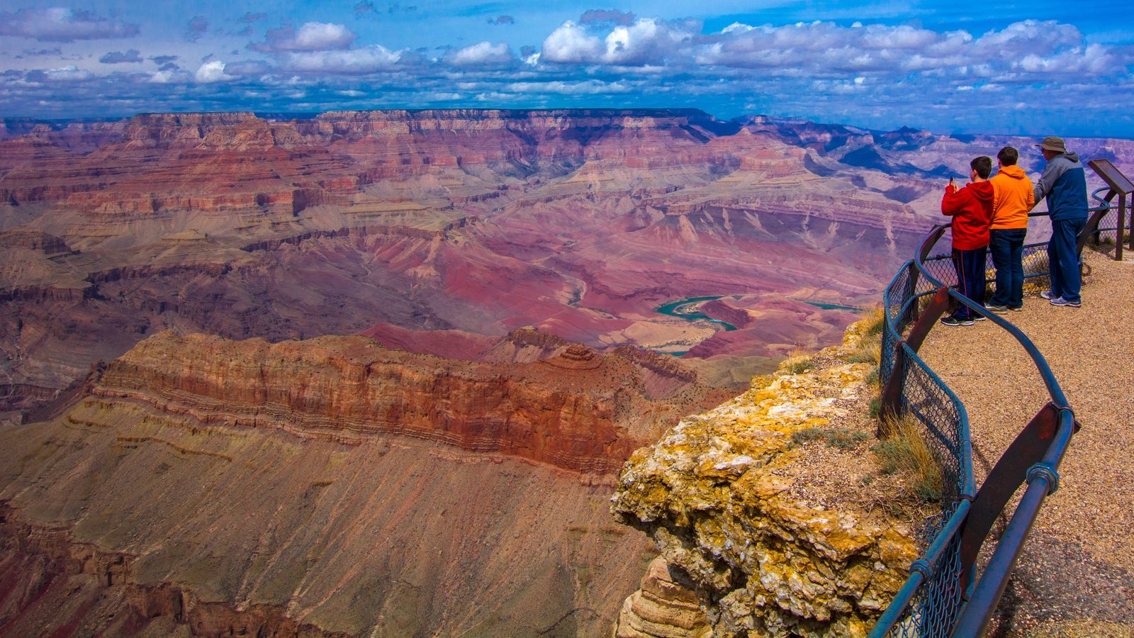 A sweeping panoramic view of the Grand Canyon from a South Rim overlook, with vibrant layered red and ochre sandstone formations and the turquoise Colorado R…