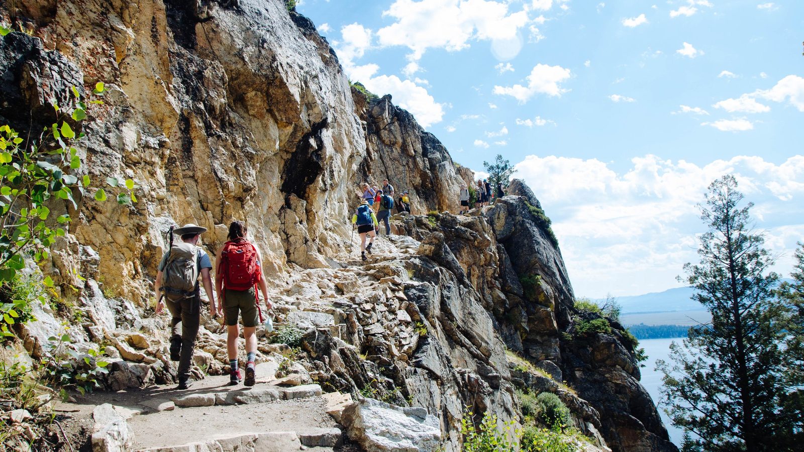 A group of hikers ascends a rocky cliff-hugging trail with stone steps carved into the mountainside, with a blue lake visible below through pine trees on the…