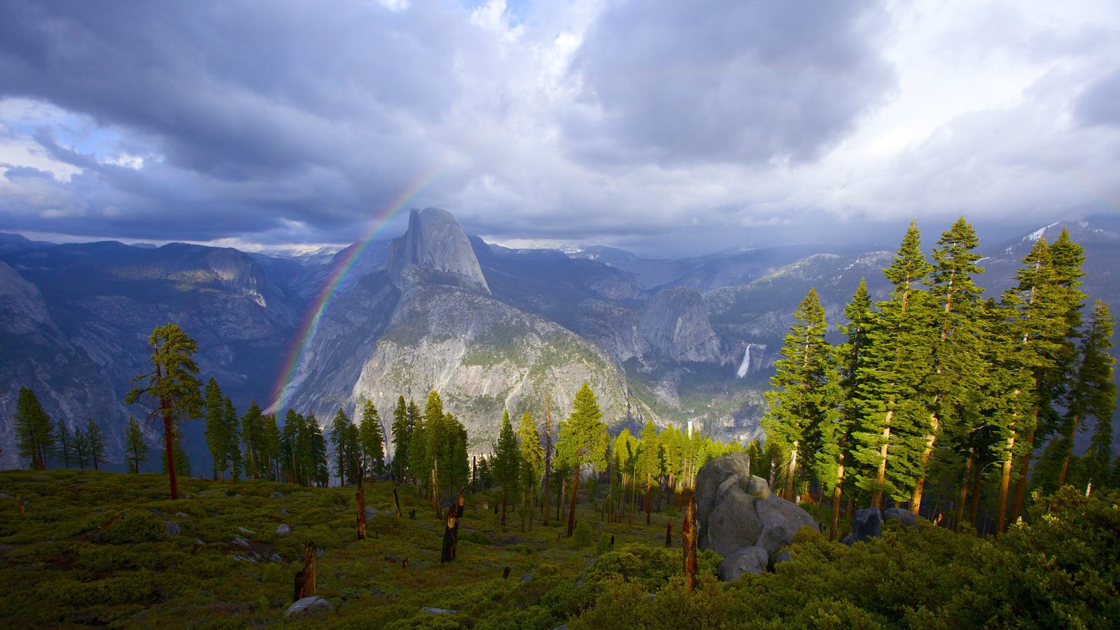 A panoramic view from Glacier Point in Yosemite National Park shows Half Dome rising prominently in the center, with a vivid rainbow arcing to its left under…