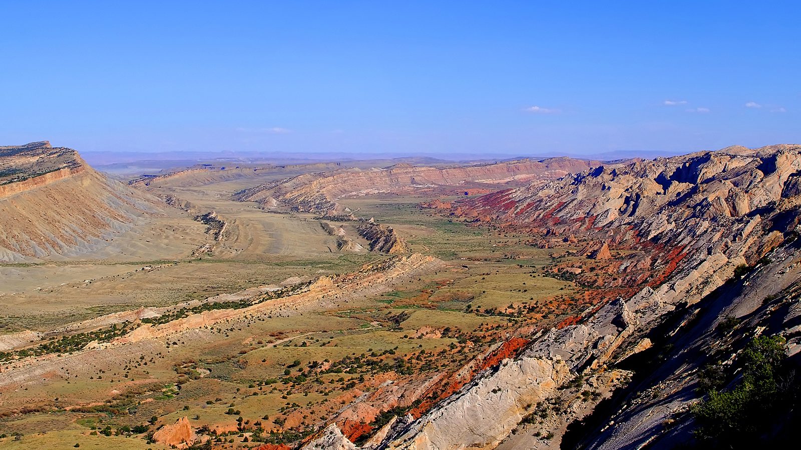 A sweeping high-elevation overlook reveals a wide valley flanked by tilted, colorful rock layers in shades of red, gray, and tan, characteristic of a Colorad…