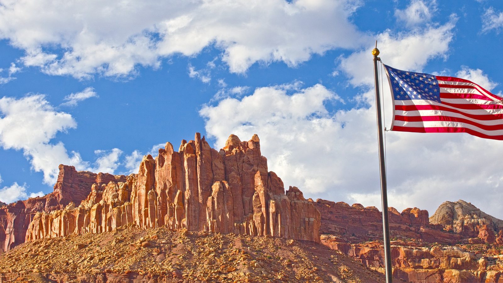 The Castle, a formation of vertical red sandstone spires at Capitol Reef National Park, rises boldly against a bright blue sky with scattered white cumulus clouds. An American flag flies in the foreground-right, bathed in warm golden sunlight enhanci