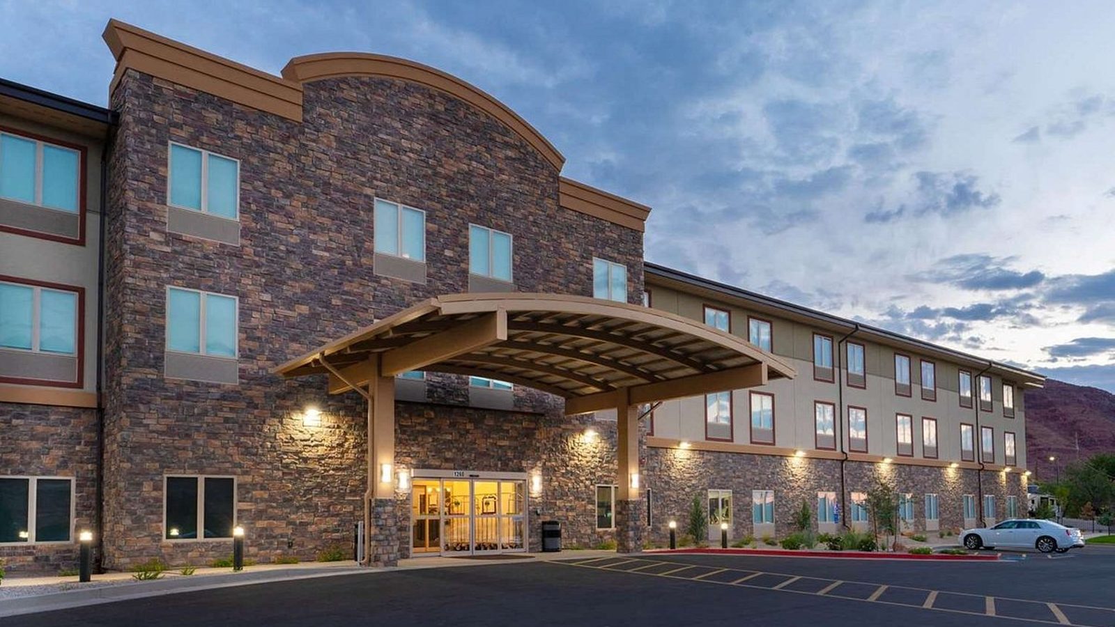 A three-story hotel or lodge building with stone facade and a covered porte-cochère entrance, photographed at dusk with warm artificial lighting against a pa…