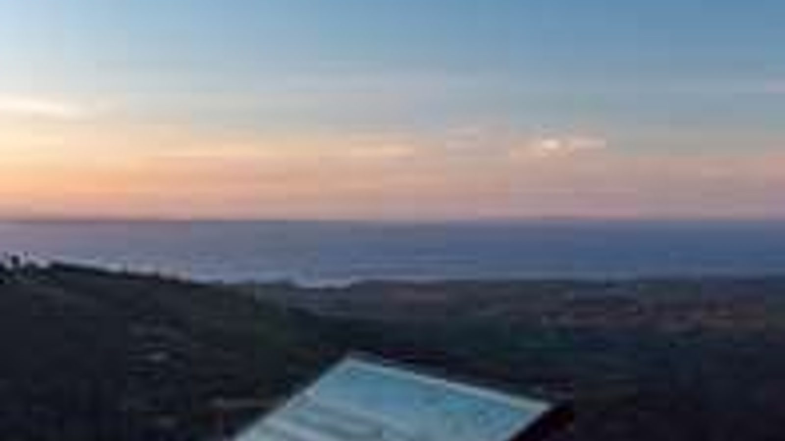 A wayside interpretive sign stands at a scenic overlook above a vast coastal or inland sea landscape at twilight, with warm sunset colors fading along the ho…