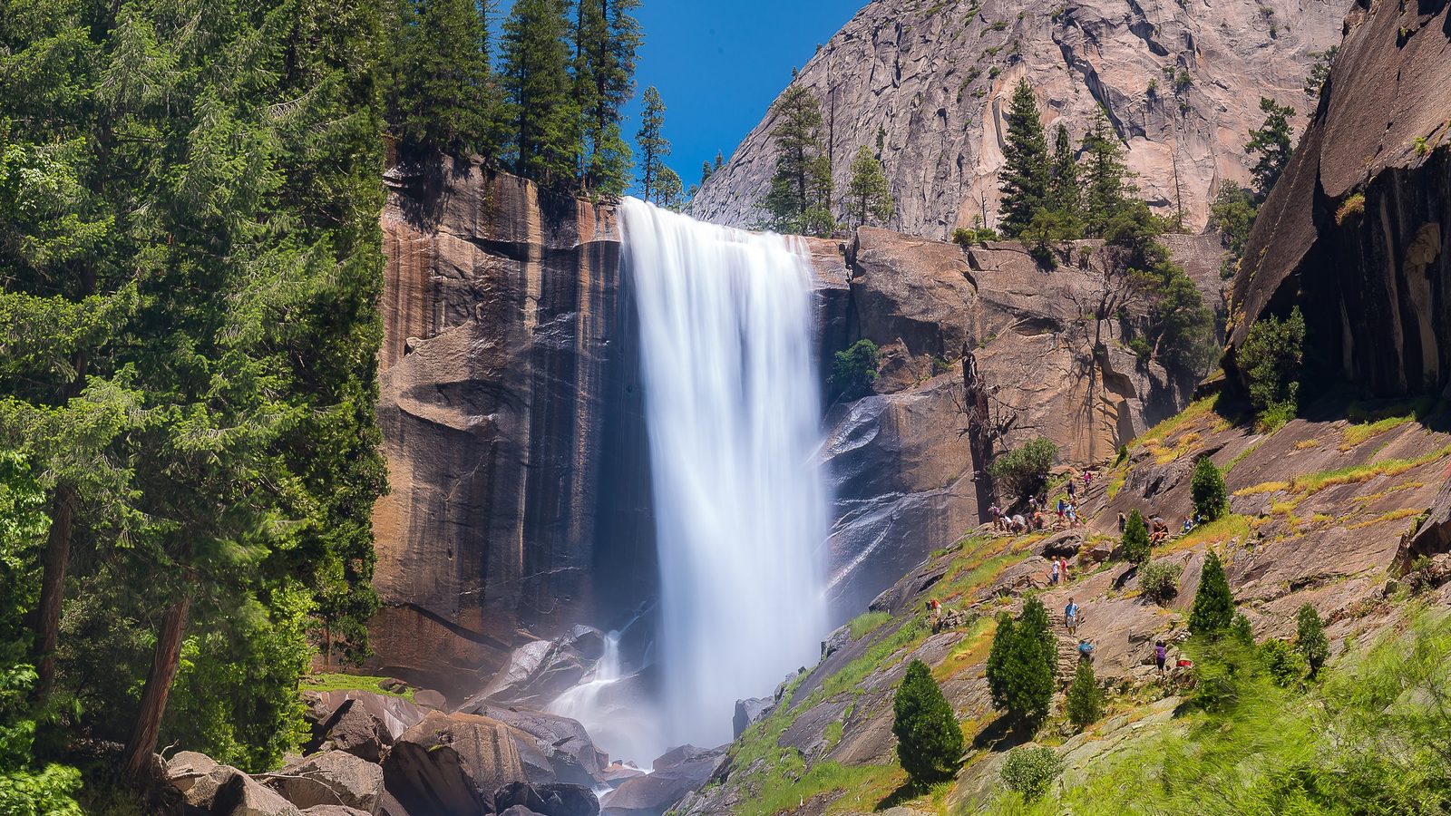 A powerful waterfall cascades down a sheer granite cliff face in Yosemite National Park, likely Vernal Fall, surrounded by lush conifer forests and rocky ter…