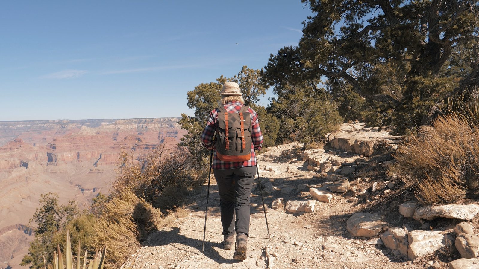 A hiker with trekking poles and a backpack walks along a rocky rim trail at the Grand Canyon, with sweeping views of the layered red sandstone canyon walls u…