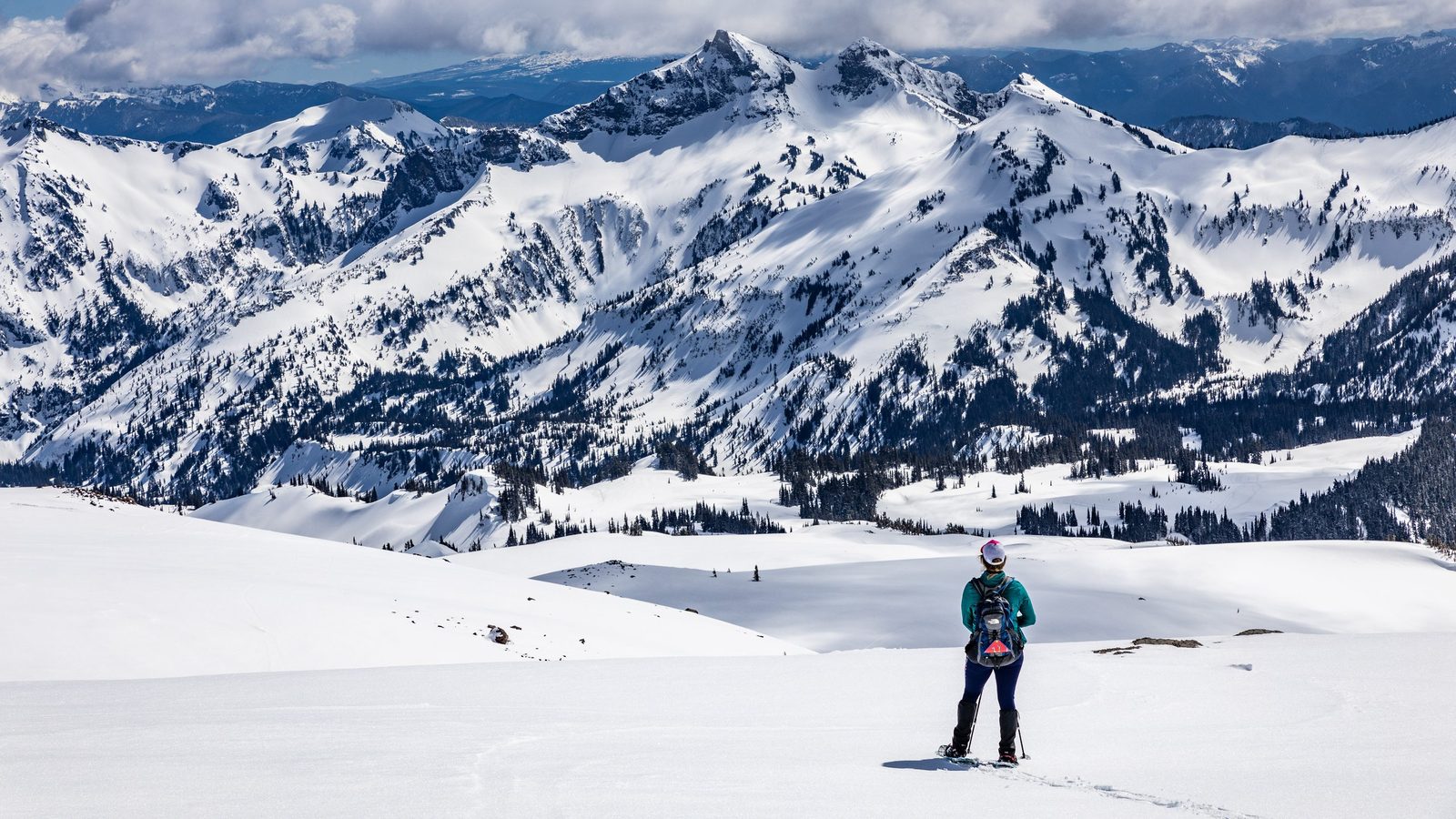A lone snowshoer stands in the foreground gazing out at a dramatic panorama of snow-covered peaks and forested valleys in what appears to be Mount Rainier Na…