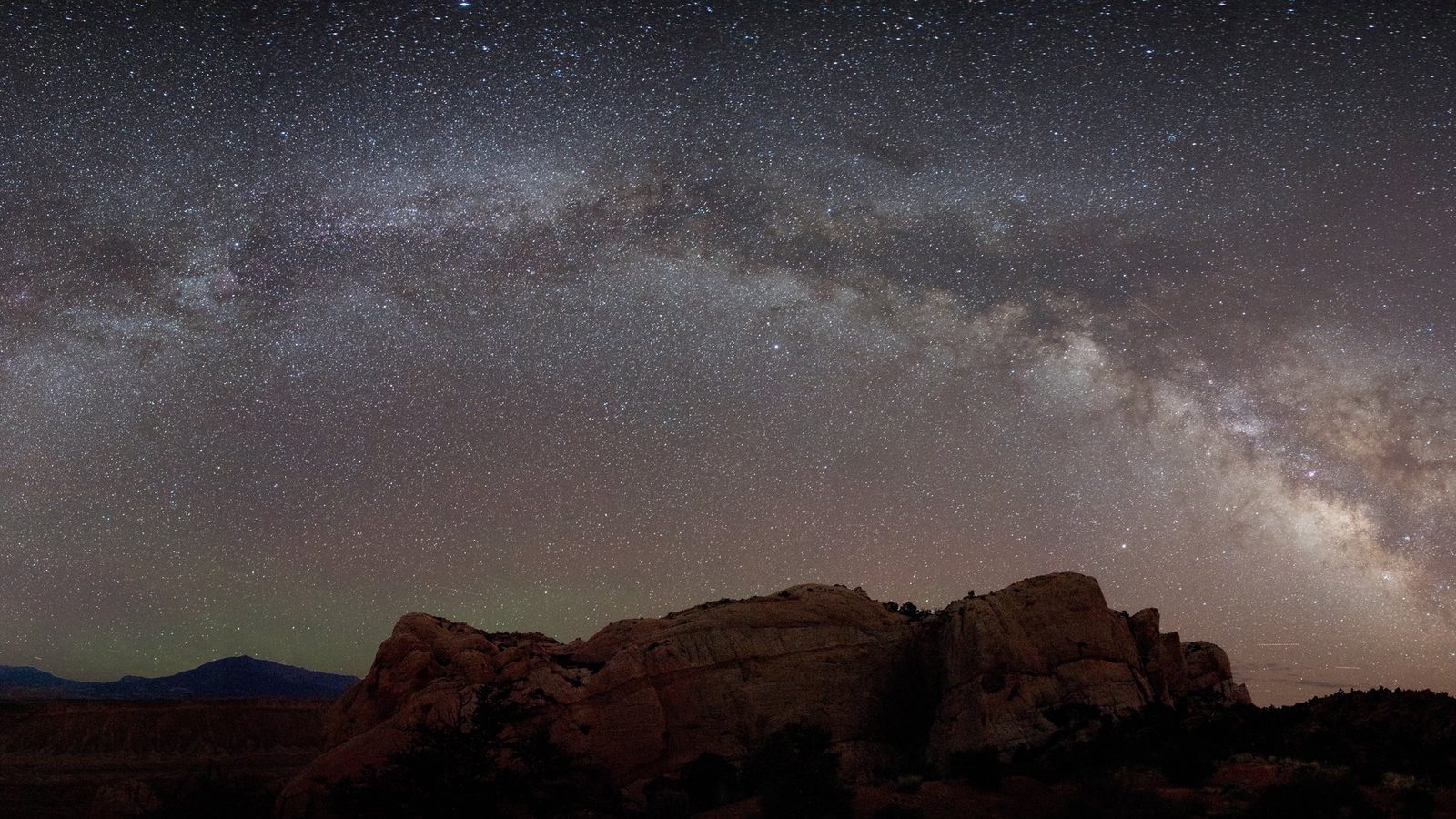 A stunning panoramic night sky scene showcasing the full arch of the Milky Way galaxy over dramatic red sandstone rock formations in a high-desert landscape.
