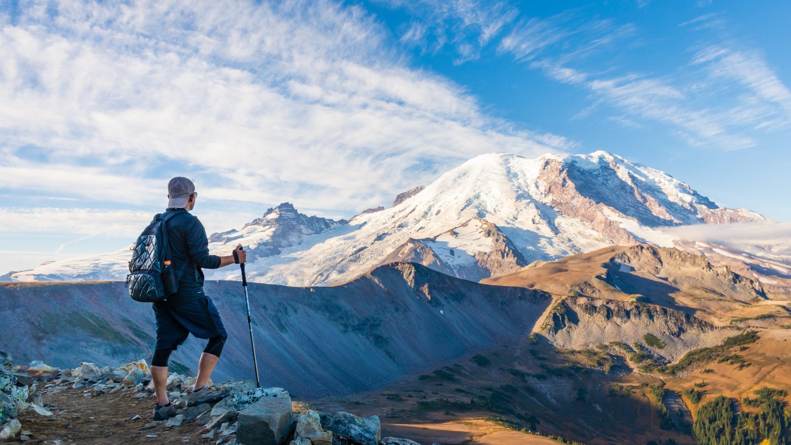 A hiker with trekking poles and a backpack stands on a rocky ridge gazing at the snow-capped summit of Mount Rainier, bathed in warm golden morning light aga…