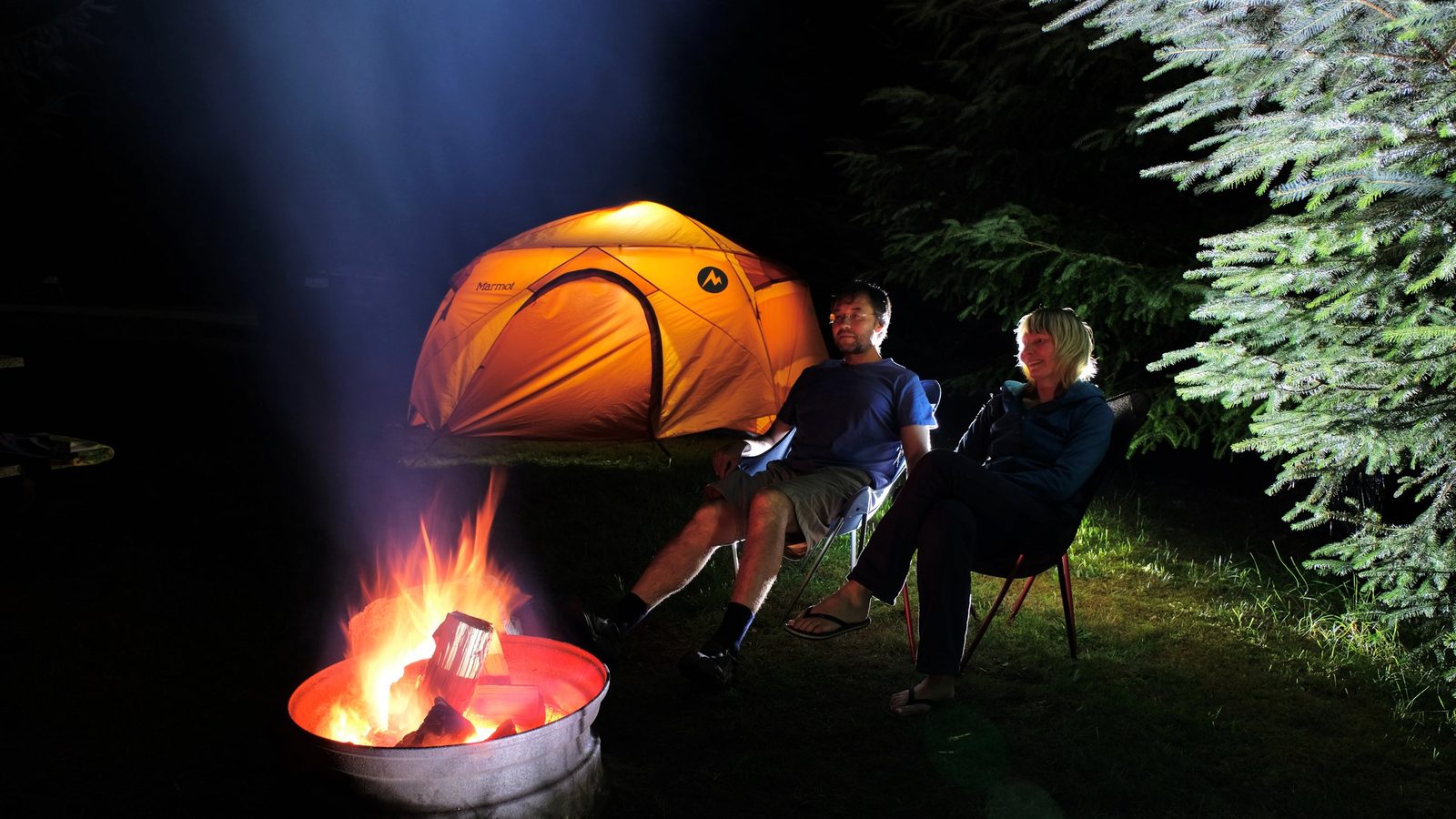 A couple relaxes in camp chairs beside a glowing orange Marmot tent and a crackling campfire in a metal fire ring, set against a dark conifer forest at night.