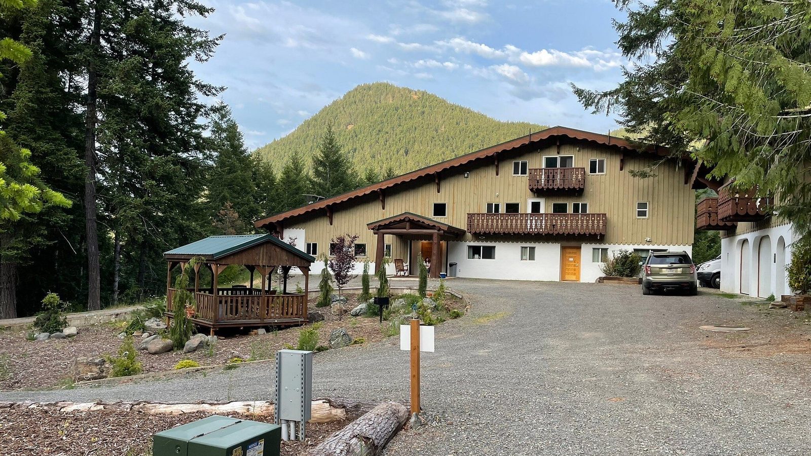 A Alpine-style lodge or inn with tan siding, dark brown trim, and decorative balconies sits at the base of a forested conical mountain peak under a partly cl…