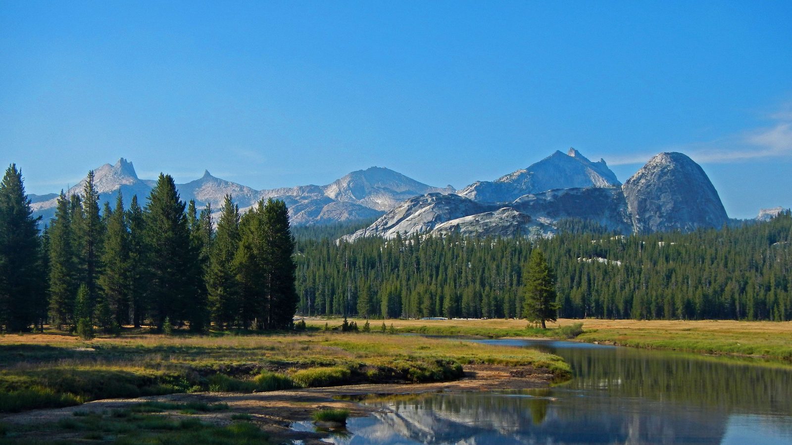 A winding river reflects granite domes and evergreen forest in an open alpine meadow, with rocky peaks rising against a clear blue sky.