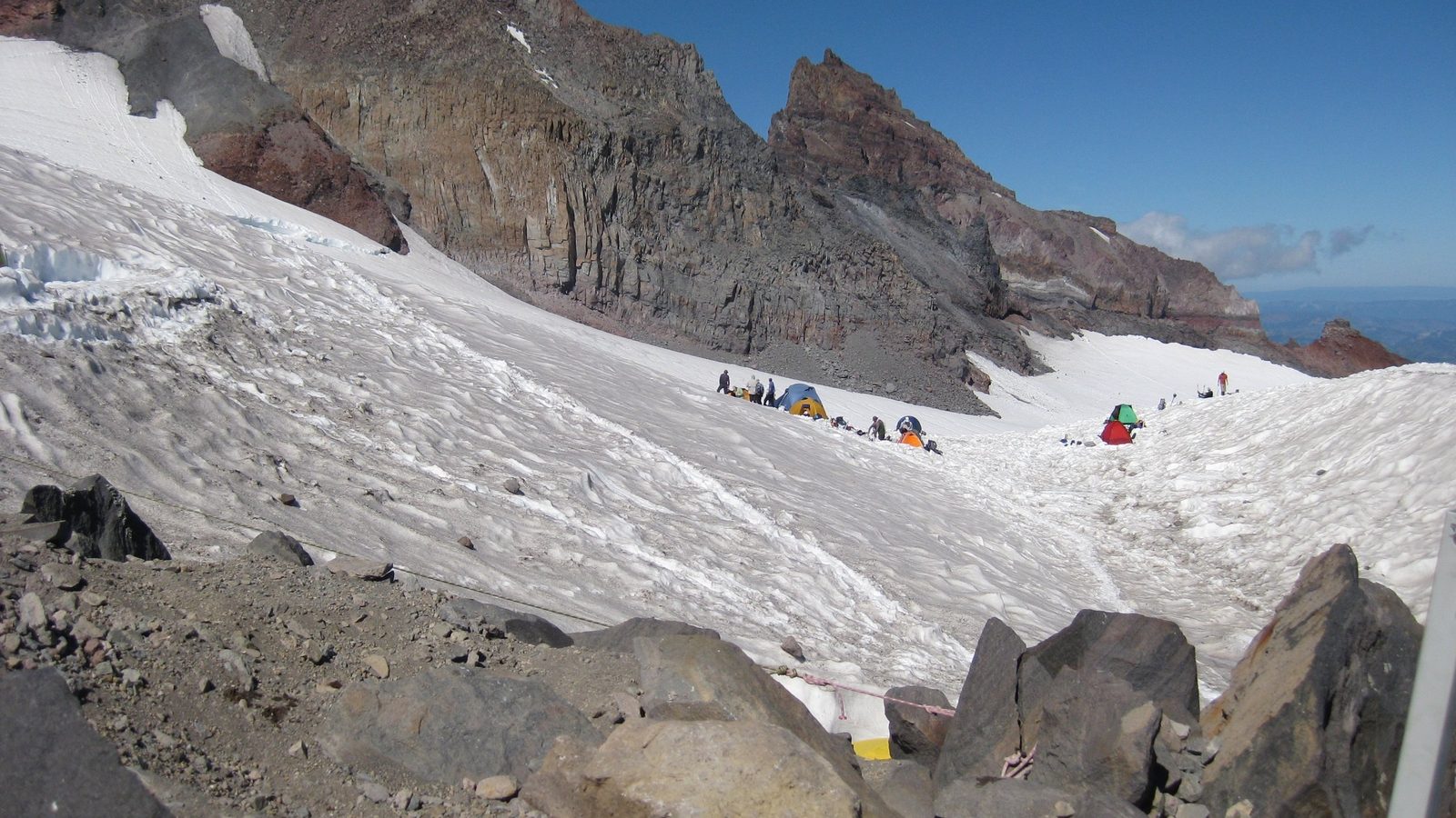 A high-altitude glacial basin on Mount Rainier shows a broad snowfield flanked by rugged volcanic rock cliffs, with several colorful mountaineering tents pit…