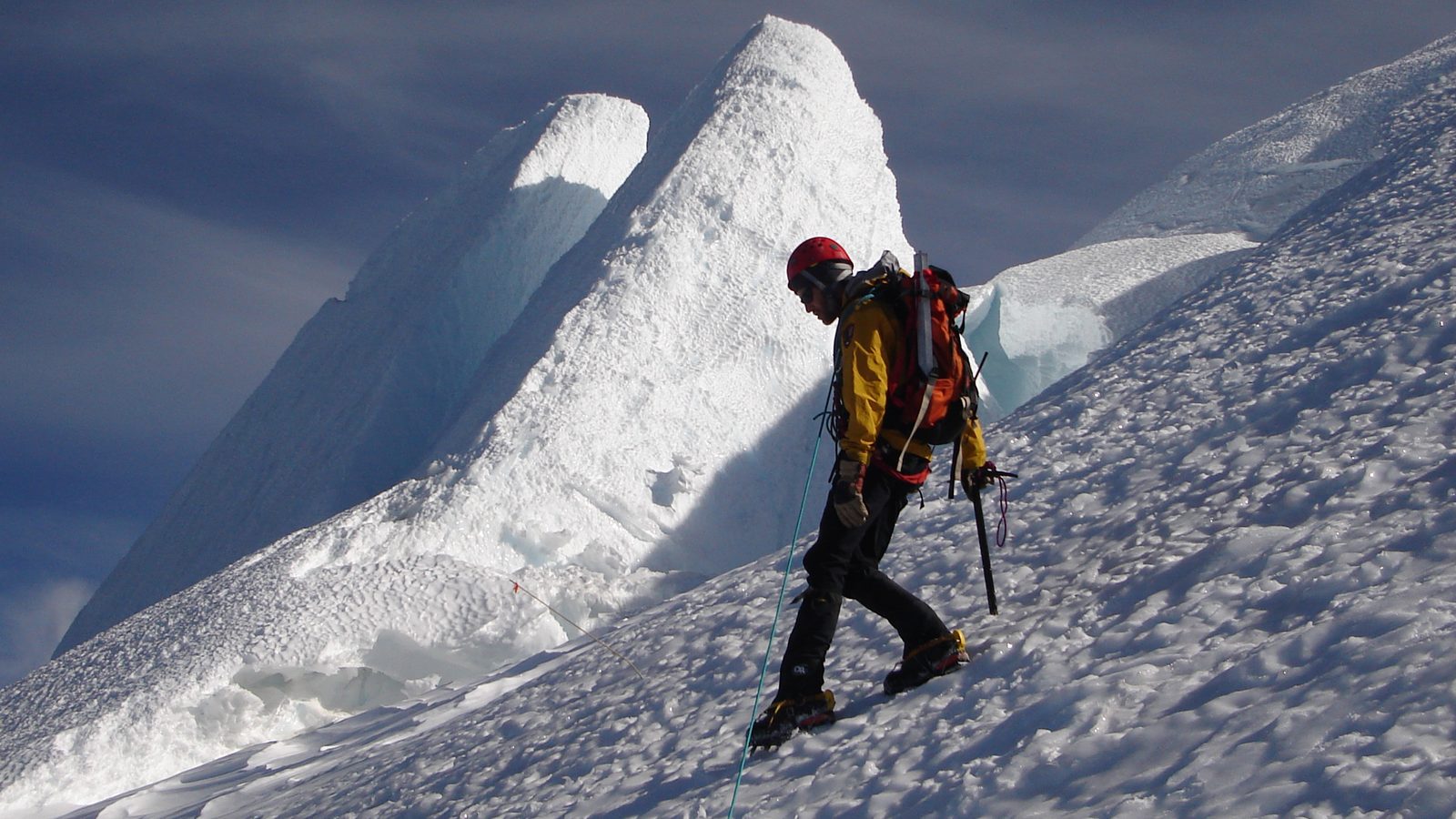 A mountaineer equipped with helmet, harness, and ice axe traverses a steep snow slope at high altitude, with large seracs and ice towers rising sharply in th…