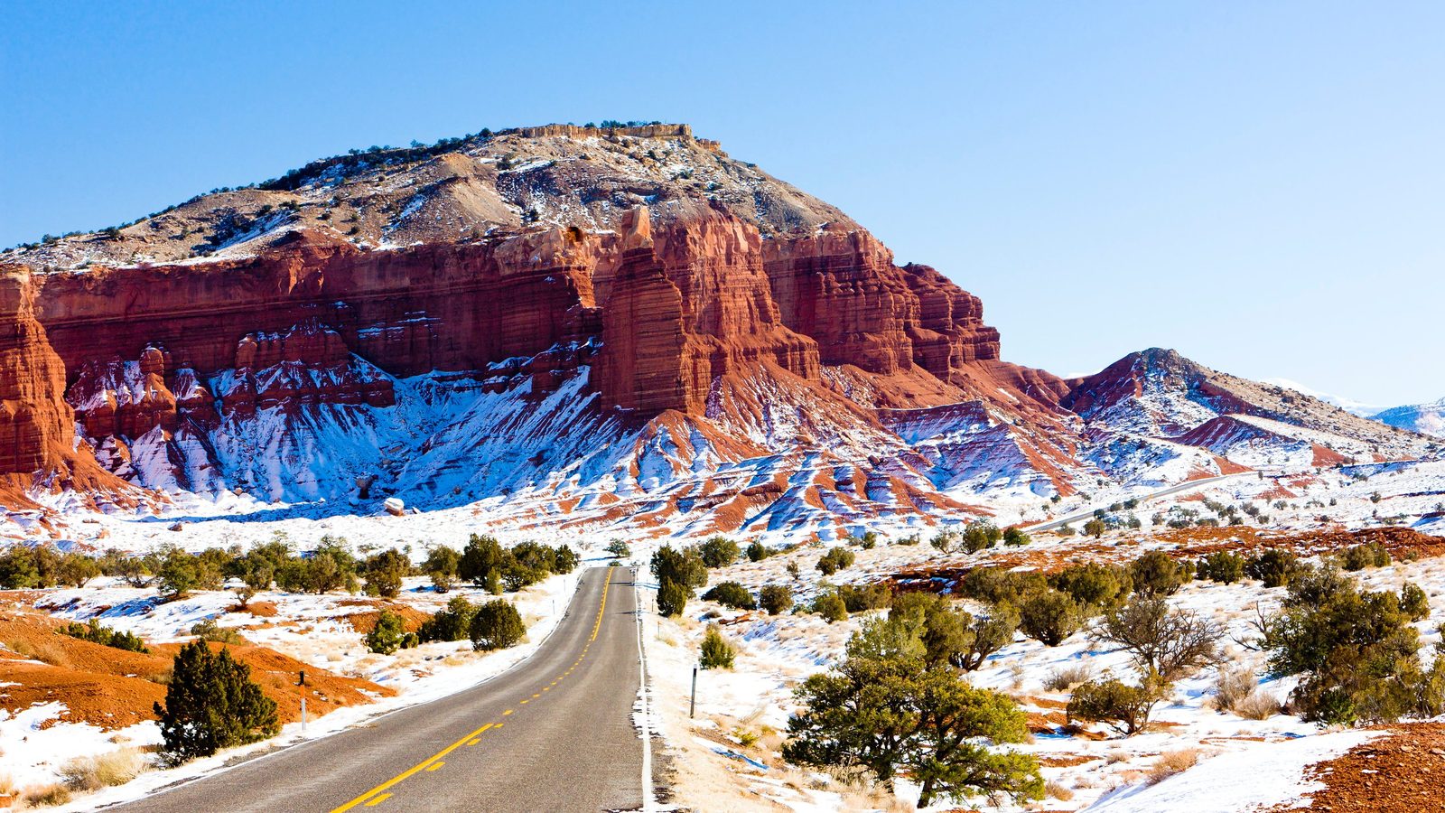 A two-lane highway cuts through a snow-dusted desert landscape toward dramatic red sandstone cliffs and buttes, bathed in crisp winter sunlight under a vivid…