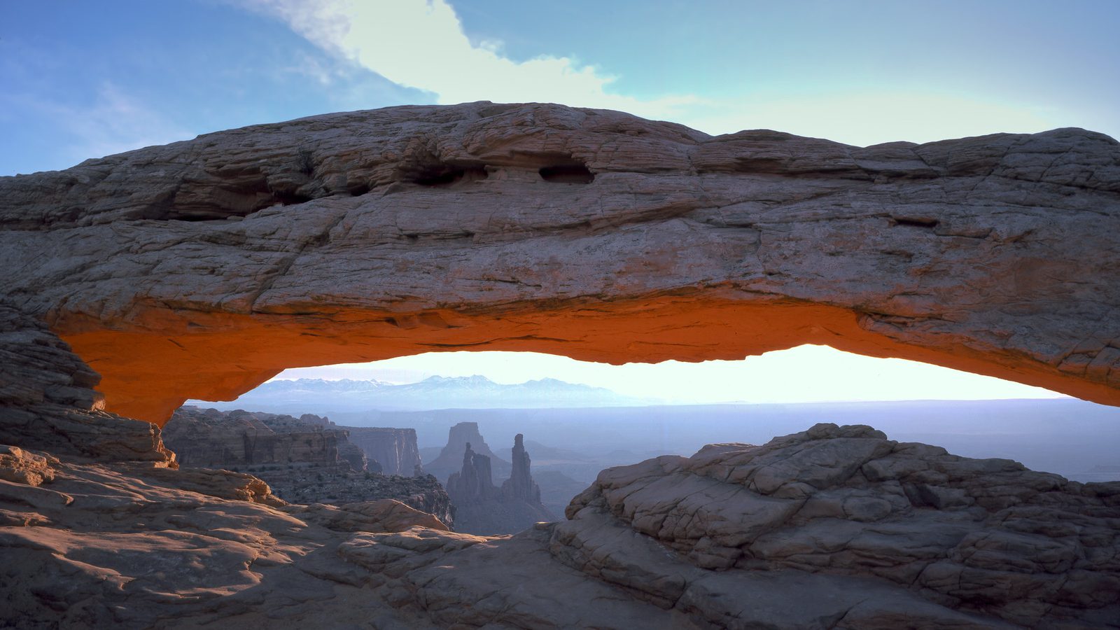 Mesa Arch at Canyonlands National Park glows with warm orange-red light reflected from sunrise on its underside, framing a vast canyon landscape with distant…