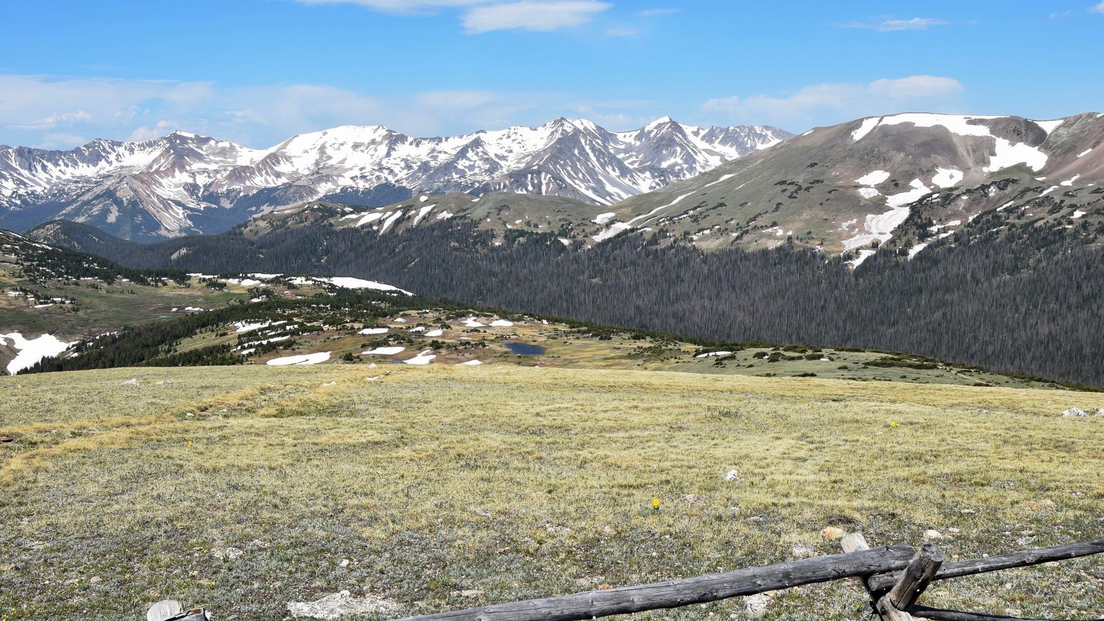 A high-alpine tundra meadow opens into a broad valley with patches of snow and a small pond, backed by a jagged range of snow-capped peaks under a bright blu…